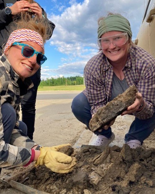 2 field techs crouching on pavement, smiling, and holding large slabs of dried mud