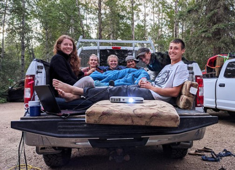 Field techs sitting in the bed of a pickup truck with cushions and sleeping bags, using a laptop and projector to watch a movie