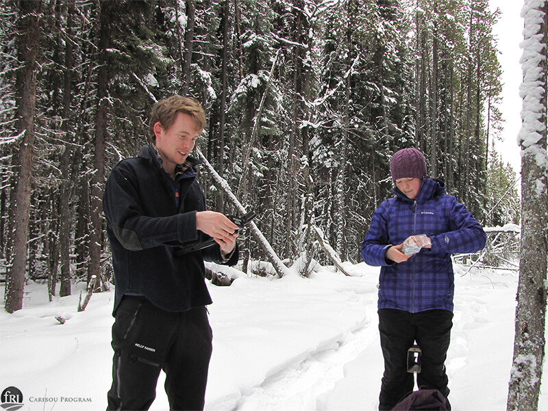 Two staff members stand beside a snowy forest to set up a trailcam.