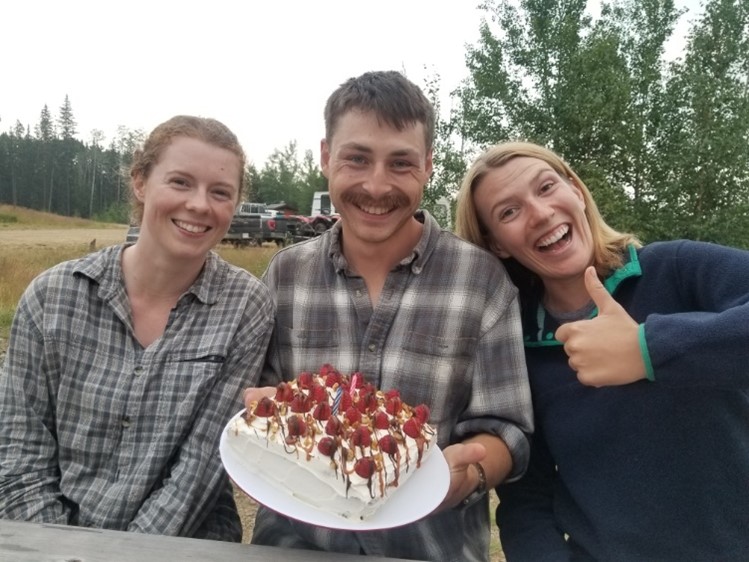 3 smiling field crew sitting at a picnic table holding a cake with berries