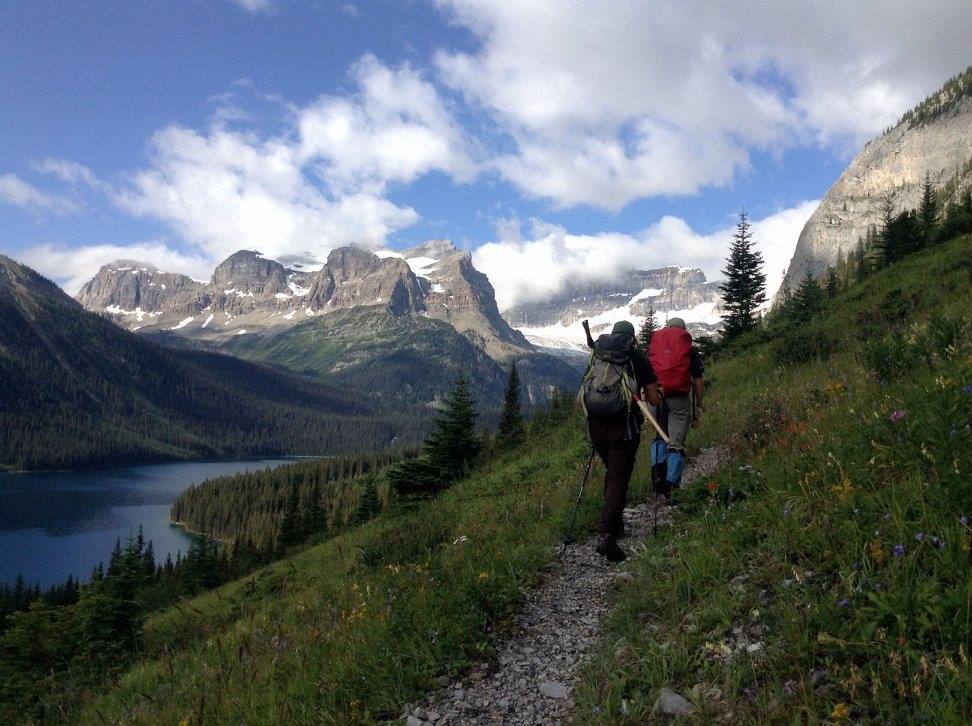 2 hikers on a trail on a slope with wildflowers passing a lake with mountains around