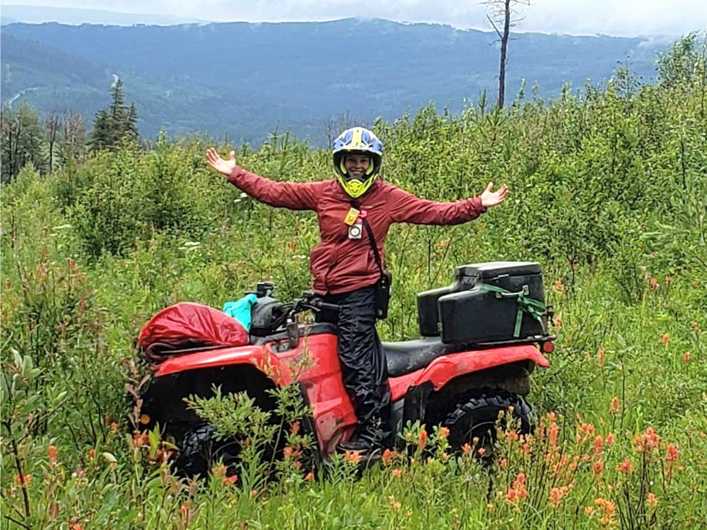 A crew member on a quad, arms wide, in a wildflower meadow with foothills in the background