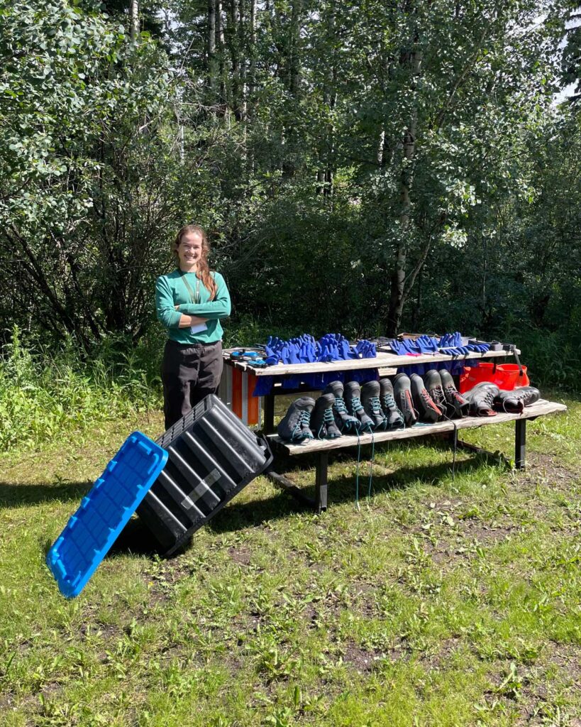 a scientist at a picnic table with many pairs of boots, gloves, and other gear