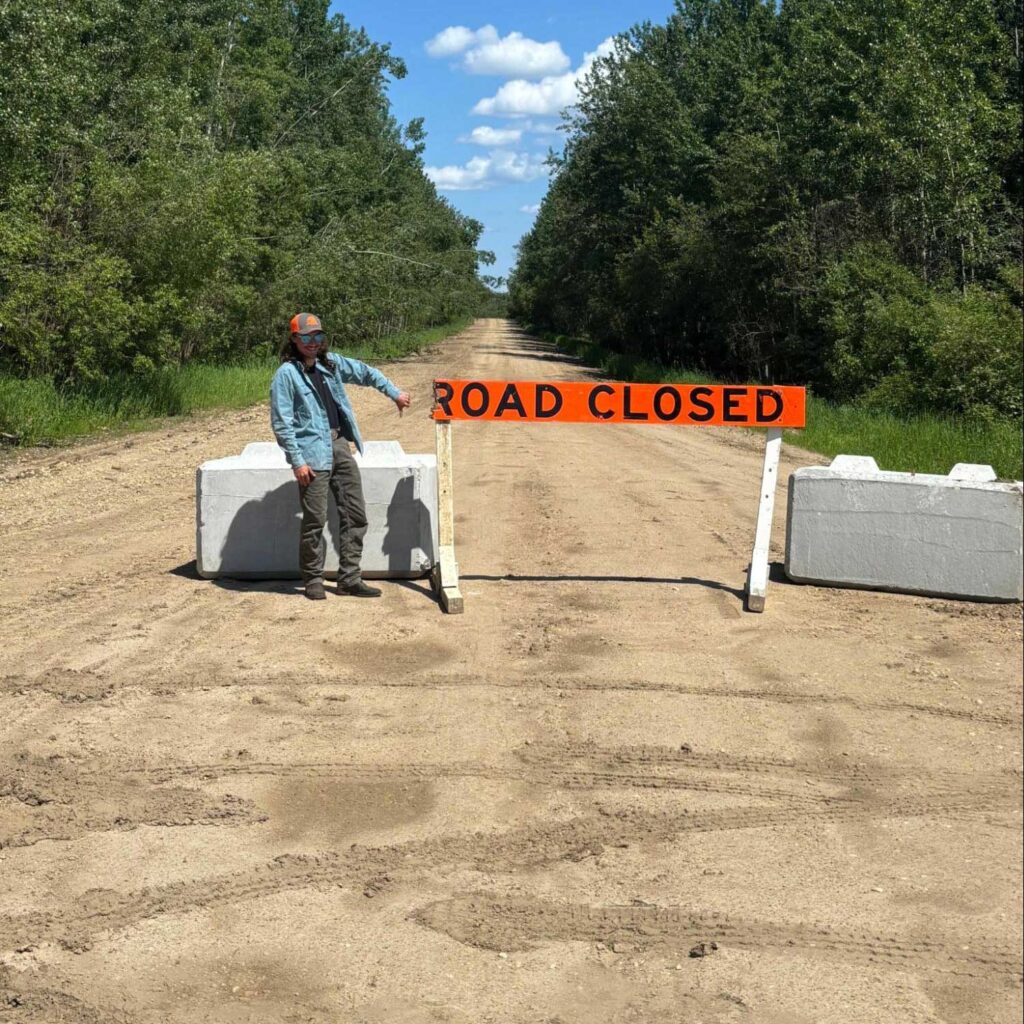 a field tech stands beside an orange road closed sign and two concrete barriers blocking a gravel road through a forest. thumbs down. photo: javier puac