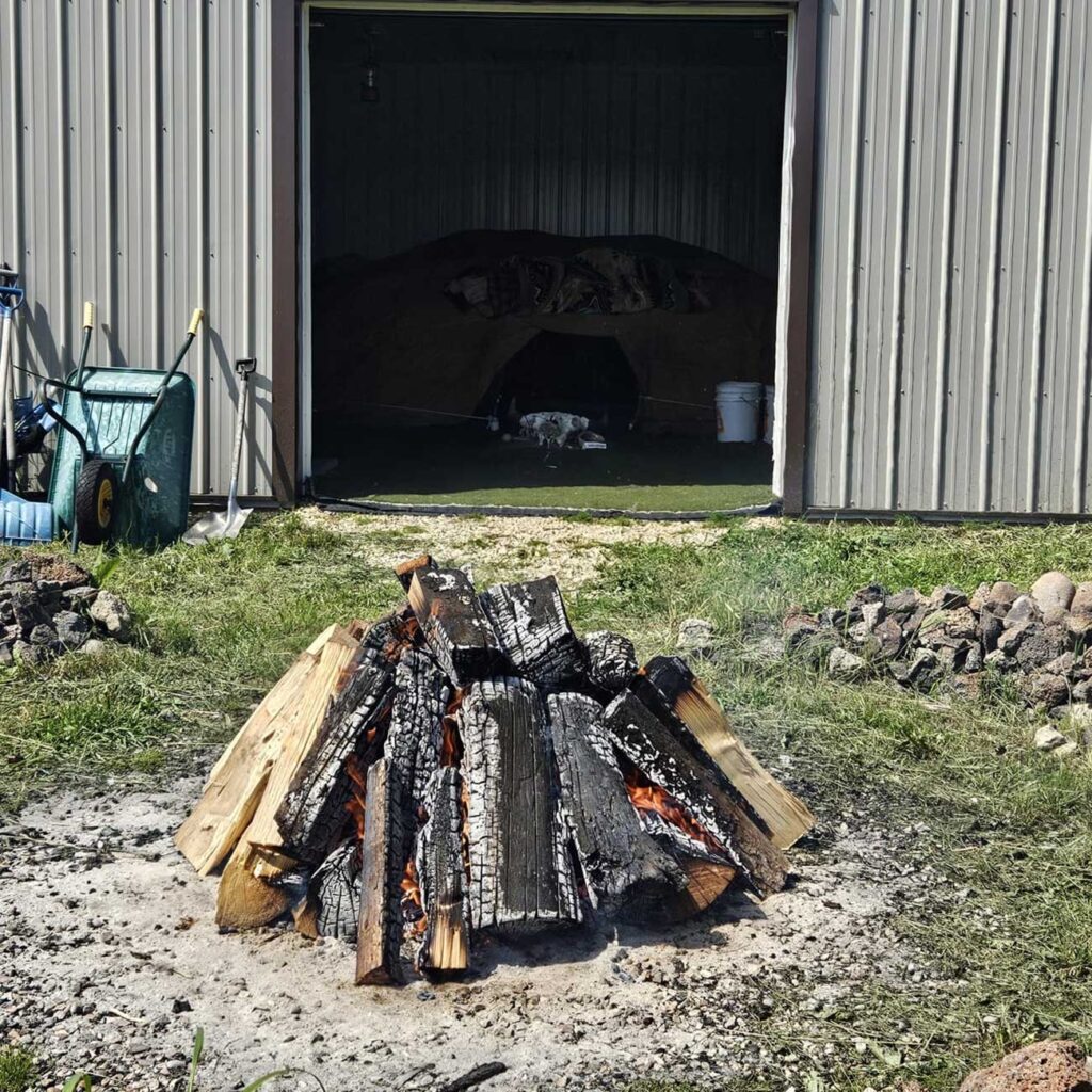 a pile of charred firewood is heaped around glowing embers in front of a large warehouse or shed with tools photo: solène williams