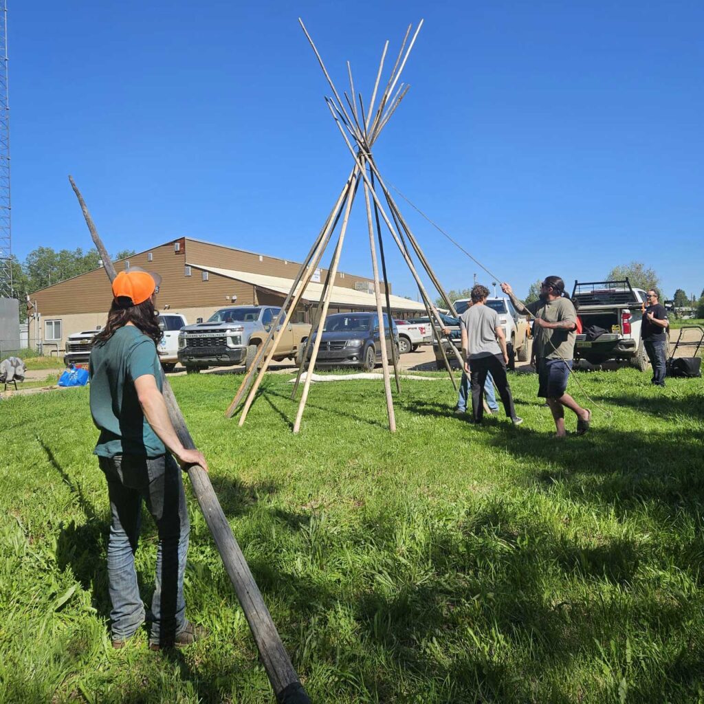 four people set up poles to create a tipi in a grassy area. trucks and a community centre in the background photo: solène williams