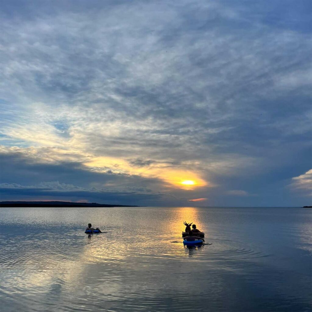two people, silhouetted against a rising sun, float in inflatable rafts on lesser slave lake. the rippling cirrus clouds in the sky mirror the surface of the lake photo: skylar shepherd.