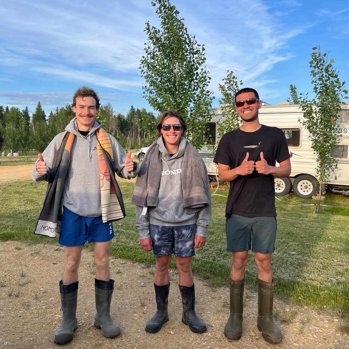 three field techs smile into the camera, wearing bathing suits, towels, and rubber boots. they are in a trailer park. photo: skylar shepherd