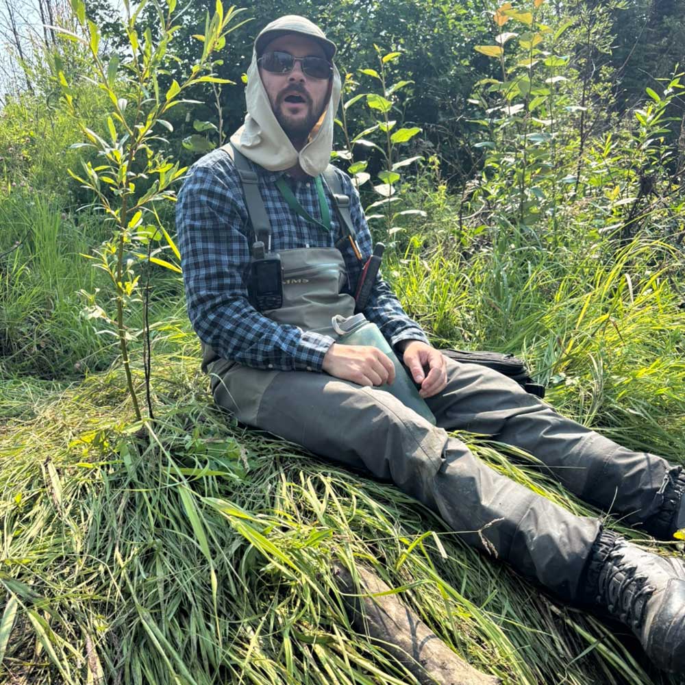 sam sits on a grassy river bank with trees around