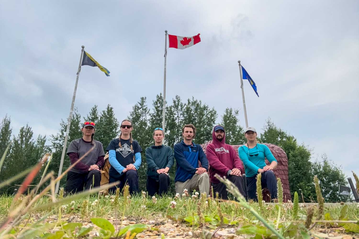 six crew members sit in the grass in front of three flags - canada, alberta, swan river